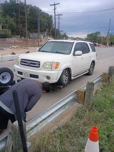 Tow truck operator in a grey sweatshirt bending down to secure the front wheel of a white Toyota SUV onto a flatbed tow truck beside a metal guardrail on a roadside in Austin, with traffic cones, utility poles, and an overcast sky in the background.
